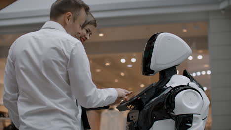 Two-people-a-man-and-a-woman-communicate-with-a-robot.-Press-the-robot-with-your-hands-on-the-screen.-A-robot-assistant-interacting-in-a-shopping-center-with-a-couple.-Robot-assistant-in-the-trading-floor